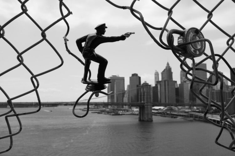 black and white close up photo of a toy man in the chain link fence on a bridge with the city skyline in the distance