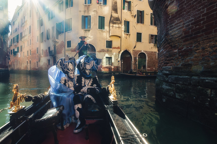 Costumed and masked characters in gondola on Venice canal passing under bridge with sunflare