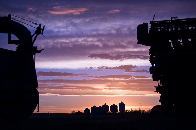 Farm buildings and equipment are shown in silhouette with the sun setting behind them